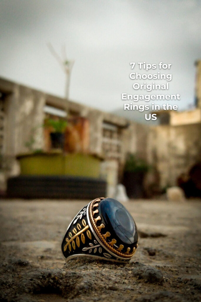 Original engagement ring with dark blue gemstone on rough ground, blurred buildings and cloudy sky behind