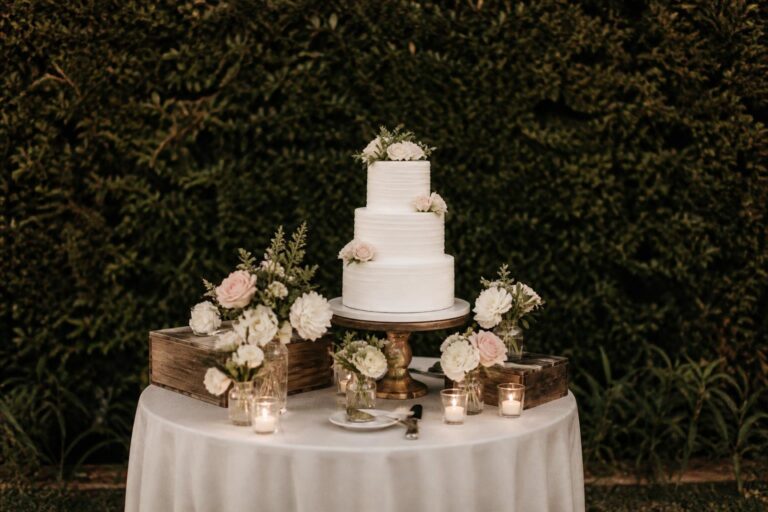 Chic wedding cake display table with tiered cake on pedestal, soft florals, greenery, and elegant linens